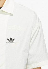 Polo bianco realizzato in tessuto morbido, con logo Adidas nero e stampa "BASKETBALL" sul lato sinistro del petto. Include un colletto classico.