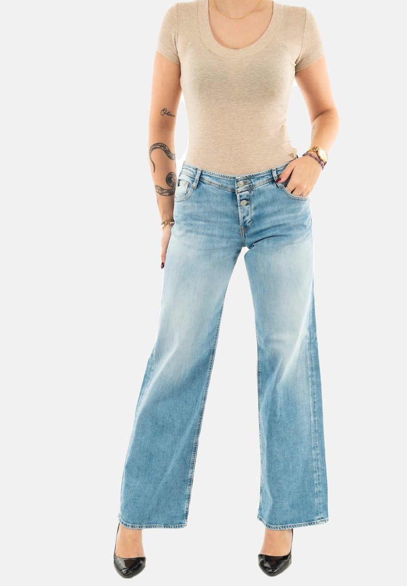 Jeans de pierna ancha color azul claro con un lavado desgastado y cierre de botones, combinados con una camiseta ajustada beige y tacones altos negros.