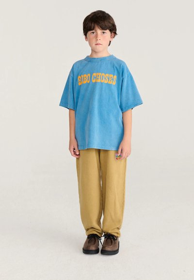 Garçon aux cheveux bruns debout portant un t-shirt bleu oversize avec un texte jaune, un pantalon moutarde et des chaussures marron sur un fond uni.