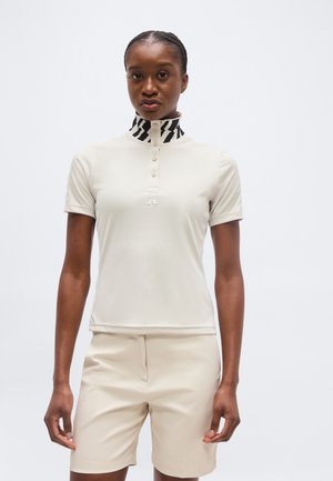 Γυναίκα που φοράει μια μπεζ κοντομάνικη πόλο μπλούζα με μαύρο και άσπρο μοτίβο στον ψηλό γιακά και ταιριαστά μπεζ σορτς, όρθια με ευθεία στάση.