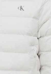 Λευκό μπουφάν puffer με ραμμένα τμήματα, ασημί λογότυπο και λεπτομέρεια τσέπης με φερμουάρ στο πλάι. Μαλακό, μονωτικό υλικό.