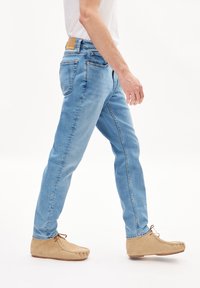 Jean en denim bleu clair avec une coupe slim, présentant une taille mi-haute, cinq poches et une patch en cuir beige sur la ceinture arrière.