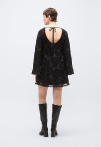 Μαύρο κεντητό φόρεμα με χαμηλή πλάτη και ζώνη, με μακριά μανίκια και ανθισμένα σχέδια· σετ με μαύρες μπότες έως το γόνατο.