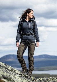 Vandrarutuställning med en grå zip-up hoodie och bruna byxor, utrustad med fickor och en ryggsäck, i en bergig landskap.