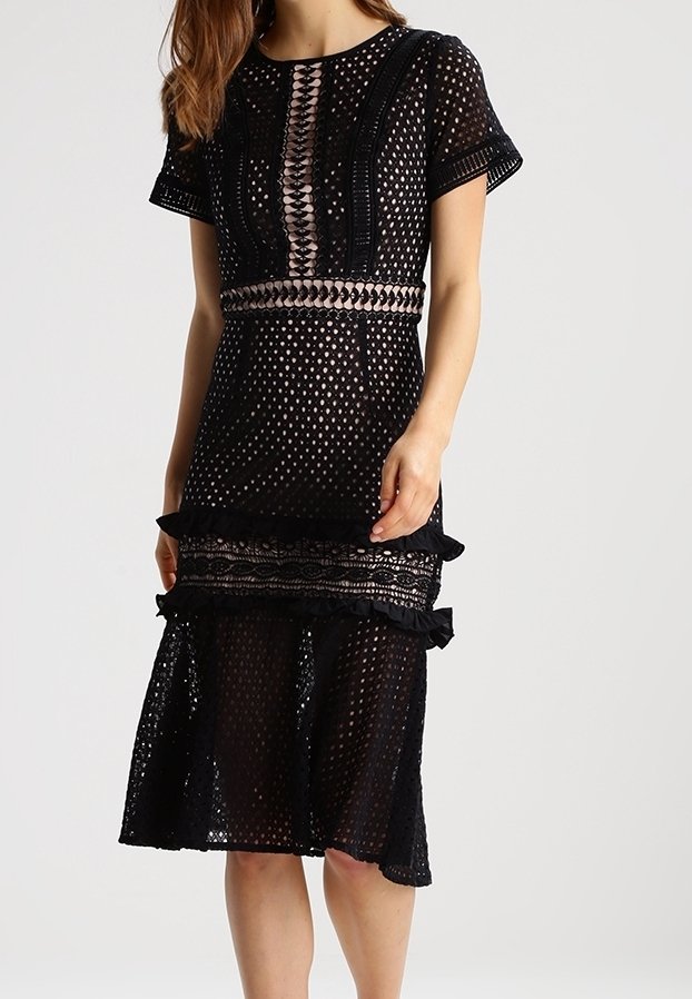 Kvinna som bär en svart spets midi-klänning med korta ärmar, geometriska utskärningar, horisontell midjeband och volangdetaljer i kjolen.