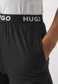 Mustat urheiluhousut, joissa on joustava vyötärö ja valkoiset "HUGO"-kirjaimet. Silkkinen pinta, oikealla puolella näkyvä sivutasku.