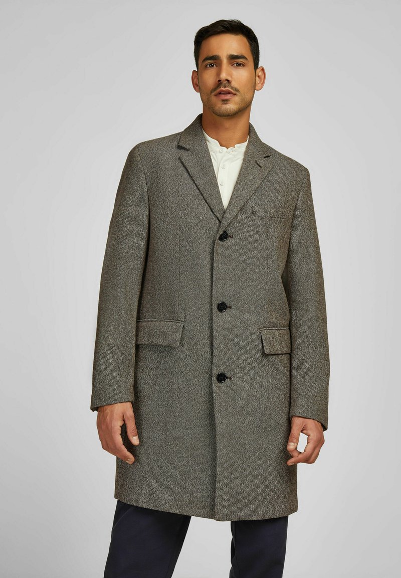 Grijze textuur wol coat met een notch-kraag, drie donkere knopen, twee voorzakken en een middellange ontwerp. Gekleed over een wit shirt.