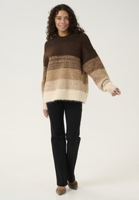 Bruine en beige gestreepte trui met een losse pasvorm, brede mouwen en een zachte, pluizige textuur. Gepaard met zwarte broeken en beige hakken.