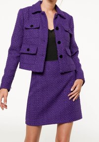 Woman wearing a textured purple jacket with black buttons and matching purple skirt, over a black top, standing against white background.
