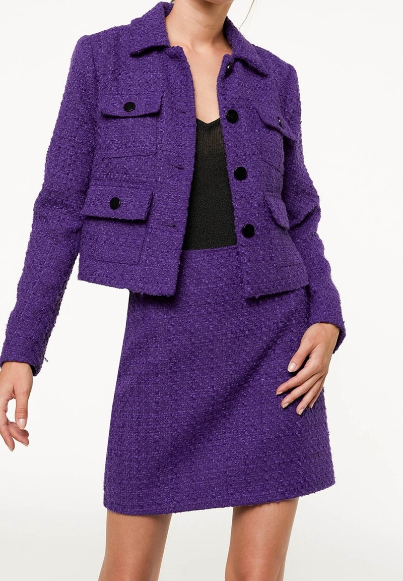 Woman wearing a textured purple jacket with black buttons and matching purple skirt, over a black top, standing against white background.