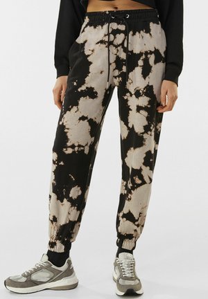 Persoon met zwart-beige tie-dye joggingbroek met elastische boorden en grijze sneakers, staand tegen een effen achtergrond.
