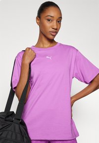 Lilla bomulds t-shirt med rund hals og korte ærmer, der har et lille hvidt Puma-logo. Modellen holder en sort taske med stropper.