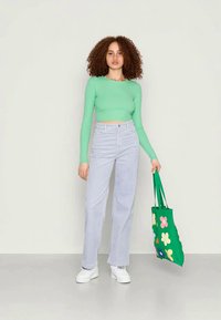 Top verde ajustado de manga longa, jeans de cintura alta azul claro, ténis brancos e uma mala tote verde com padrões de flores multicoloridas.