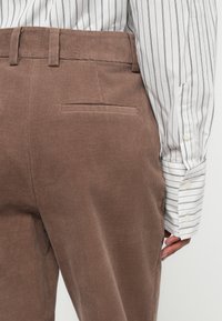 Persoon in een bruine corduroy broek en een wit shirt met lange mouwen en zwarte strepen, hand rustend naast de achterzak van de broek.