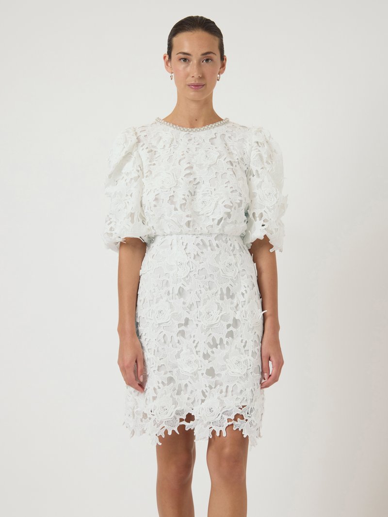 Λευκό δαντελένιο φόρεμα με λουλουδάτο σχέδιο, με φουσκωτά κοντά μανίκια και εφαρμοστή μέση. Η κόψη είναι περίτεχνα σχεδιασμένη με καμπυλωτές άκρες.