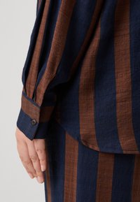 Ruda ir tamsiai mėlyna dryžuota palaidinė su tekstūruotu audiniu, pasižyminti sagčių apykakle ir pritaikytu siluetu. Detalus rankovės ir rankos vaizdas.