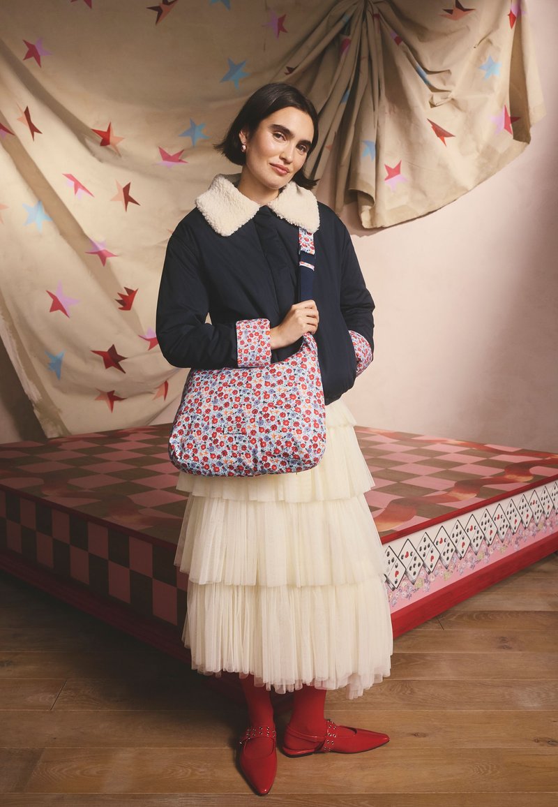 Giacca imbottita blu navy con colletto in pelliccia beige, borsa con motivo floreale, gonna a balze in tulle bianco e ballerine rosse con dettagli metallici.