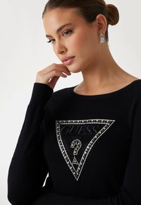 Schwarzer Pullover mit einem dreieckigen Logo, verziert mit Strasssteinen, aus strukturiertem Stoff und mit weitem Halsausschnitt. Nahaufnahme eines Modells.