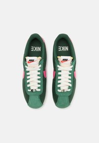 Tenisky Nike s vrškem z zelené látky, růžovými detaily, bílými tkaničkami a polstrovaným jazykem s logem Nike v černé a červené barvě.