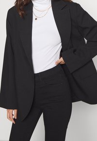 Svart skräddarsydd kavaj med öppen front, strukturerade axlar och långa ärmar; bärs över en vit polotröja och svarta jeans.