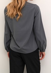 Blouse grise à manches longues avec légère brillance, coupe décontractée, manches froncées et fermeture par bouton au poignet. Portée avec un pantalon noir.