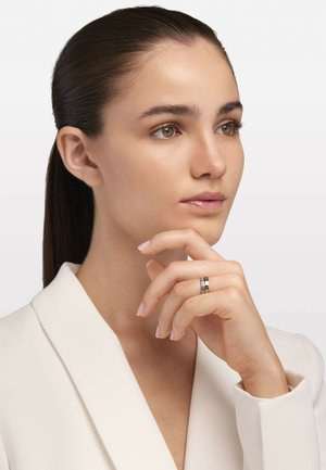 Silber- und Goldring mit einem horizontalen Band aus Diamanten, getragen an einer Hand mit hellrosa Nägeln, vor einem weißen Hintergrund.