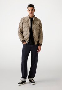 Veste bomber beige avec des détails à motifs, jean noir et baskets noires avec des accents blancs. La veste présente un col montant et une fermeture éclair.