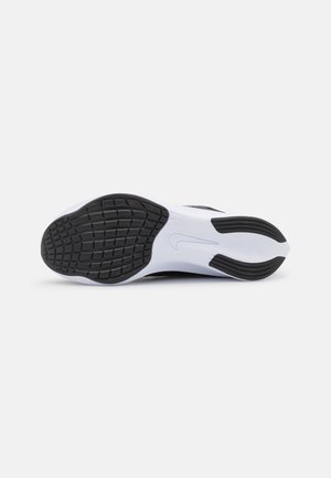 Semelle d'une basket blanche avec des patins traction en caoutchouc noirs et un logo Nike, montrée à plat sur un fond gris clair.