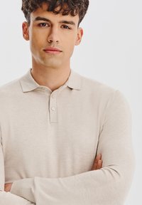 Młody mężczyzna z kręconymi brązowymi włosami, ubrany w beżową koszulę polo z długim rękawem, stoi z założonymi rękami na tle jasnego, jednolitego tła.