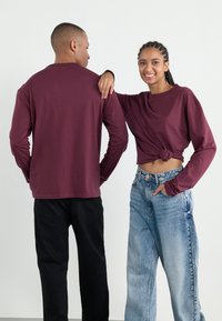 Mørkerød langermet skjorte, knyttet i midjen, sammen med lys blå high-waisted jeans. Svart bukse på den mannlige figuren i bakvinkel.