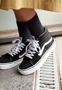 Persona che indossa sneakers alte Vans bianche e nere con lacci bianchi, calze nere e una gonna marrone, in piedi vicino a una grata metallica.