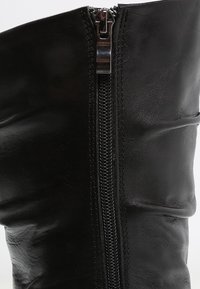 Botte en cuir noir avec une fermeture éclair verticale à l'avant, dotée de surpiqûres et d'une texture lisse et mate.
