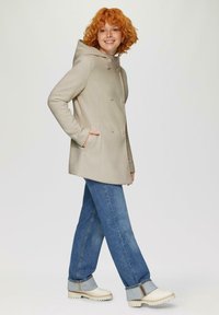 Beige gewatteerde jas met dubbelzijdig ontwerp, twee voorzakken en beige knopen. Gecombineerd met blauwe jeans en witte enkellaarsjes.