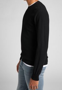 Schwarzer gestrickter Pullover mit Rundhalsausschnitt, gerippten Bündchen und Saum, der ein strukturiertes Muster aufweist. Über einem weißen Hemd mit hellblauen Jeans getragen.