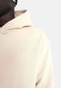 Sudadera beige con una textura suave, que cuenta con capucha ajustable con cordones, dos botones a presión y un logo bordado en el pecho.