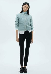 Cardigan blu chiaro, con una texture, dotato di due tasche frontali, abbinato a una camicia bianca, pantaloni neri attillati e scarpe nere.