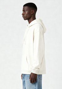 Krēmkrāsas kapucis, kas izgatavots no mīksta auduma, ar kenguru kabatu, saņemošām auklām apkaklē un ribotām aprocēm. Valkāts ar gaiši zilām džinsiem.