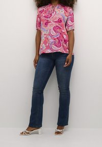 Blusa a motivi paisley rosa con maniche a sbuffo corte, scollatura arricciata, abbinata a jeans blu scuro e sandali con tacco bianchi.