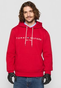 Felpa rossa in cotone con tasca a marsupio, cappuccio con coulisse e logo frontale. Abbinata a jeans blu e guanti neri.