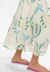 Faltenmaxikleid in hellem Creme mit grünen und pastellfarbenen Blattmustern; kombiniert mit pinken Flip-Flops. Glattes Material mit fließendem Design.