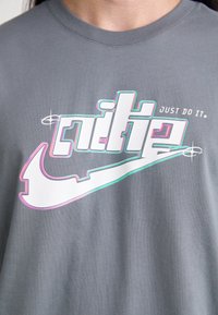 Šedé bavlněné tričko s velkým bílým logem Nike a stylizovaným textem. Design obsahuje duhové akcenty a frázi "JUST DO IT" nad logem.