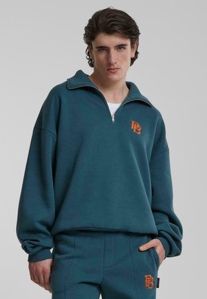 Blauwgroen, met rits gesloten pullover met een hoge kraag en oranje logo. Beschikt over losse mouwen en geribbelde manchetten. Gepaard met bijpassende broeken.