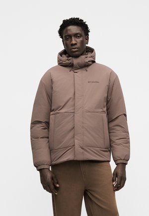 Junger Mann, der eine braune Columbia-isolierte Jacke mit hohem Kragen und Kapuze trägt, steht vor einem schlichten hellen Hintergrund.