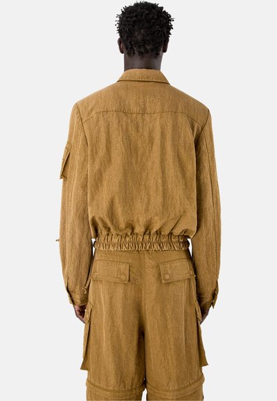 Hombre con chaqueta marrón de textura y pantalones cortos cargo a juego, mostrado de espaldas contra un fondo liso.