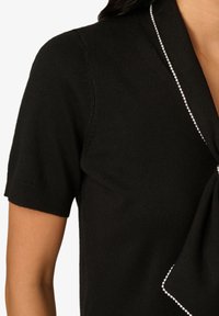 Schwarzes, kurzärmeliges Strickoberteil mit einem kragenförmigen Design, das eine strukturierte Oberfläche und weiße Verzierungen entlang des Kragens sowie eine Schleifendetail aufweist.