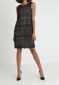 Robe en dentelle noire sans manches, avec des motifs superposés et des bords festonnés. Associée à des escarpins noirs.