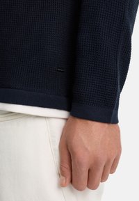 Mørkeblå strikket sweater med struktureret mønster, ribbede ærmeender og et diskret logo. Deltager iført den over en hvid skjorte, med en hånd synlig.
