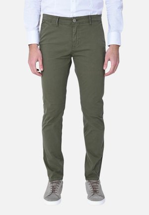 Uomo che indossa pantaloni slim verde oliva, camicia bianca abbottonata e sneaker grigie, in piedi davanti a uno sfondo bianco.