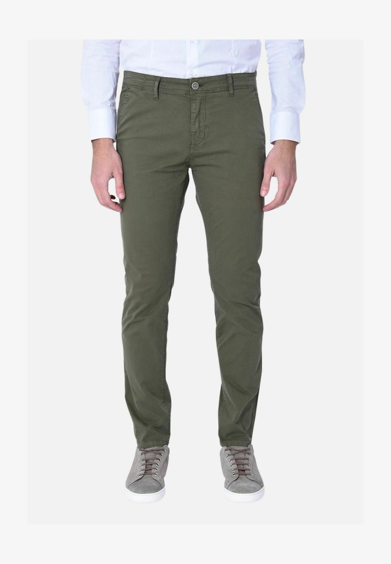 Uomo che indossa pantaloni slim verde oliva, camicia bianca abbottonata e sneaker grigie, in piedi davanti a uno sfondo bianco.