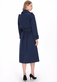 Marineblauwe trenchcoat met een getailleerde taille, lange mouwen, dubbele knoopsluiting aan de voorkant en schouderepauletten. Gladde stof met een lengte tot halverwege de kuit.